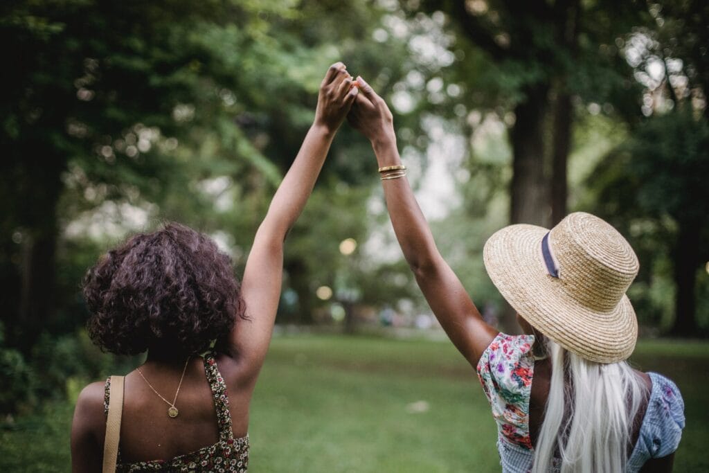 two black women hold raised hands, view is from the back. They are outdoors. One wears a hat, the other has black curly hair.