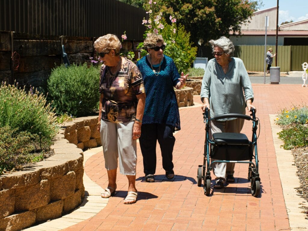 three elderly women walking on a paved pathway near a garden, one woman uses a walker.