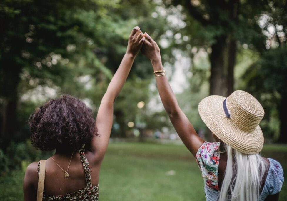 two black women hold raised hands, view is from the back. They are outdoors. One wears a hat, the other has black curly hair.