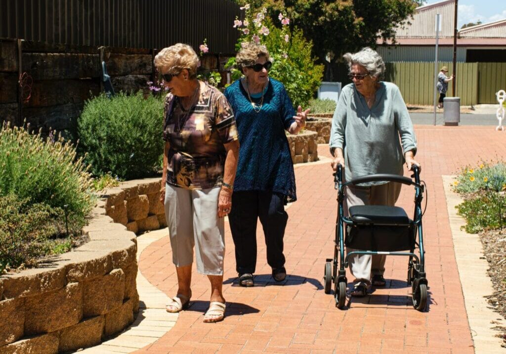 three elderly women walking on a paved pathway near a garden, one woman uses a walker.